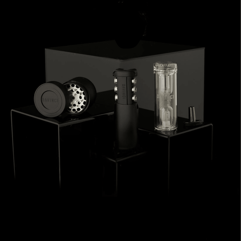 Three DaVinci accessories displayed on glossy black acrylic plinths against a dark background. From left to right: a matte black herb grinder shown open to reveal its perforated grinding chamber, a tall matte black vaporiser with silver accent buttons and 'DAVINCI' engraved on the side, and a clear hydrotube with the DaVinci logo etched into it, accompanied by a small black mouthpiece adapter.